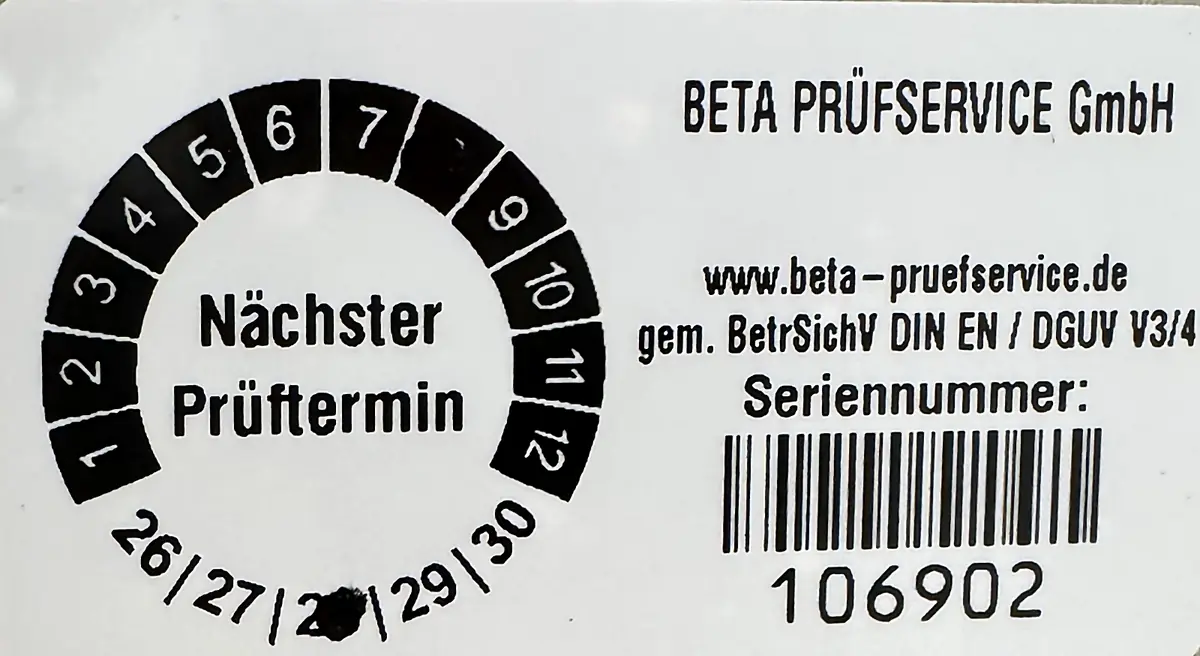 Prüfplakette der Beta Prüfservice GmbH auf einem geprüften Gerät. Die Lochung im Ring markiert Monat 8, die Jahreszahl 28 ist durchstanzt: nächster Prüftermin 08/2028. Rechts stehen die Internetadresse, die Angabe „gem. BetrSichV DIN EN / DGUV V3/4“ sowie Barcode und Seriennummer 106902.