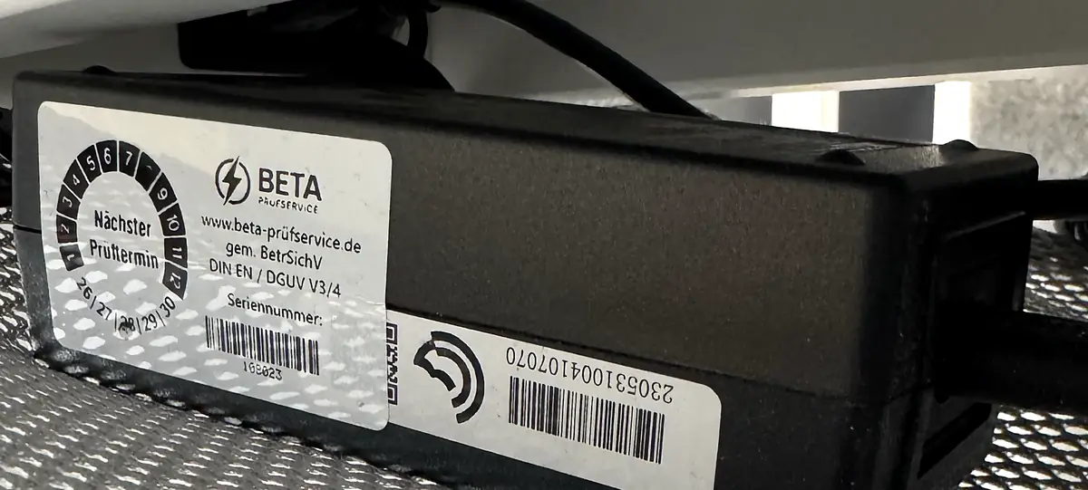 Ein Netzteil unter einem Arbeitsplatz, versehen mit der Prüfplakette der Beta Prüfservice GmbH. Die Plakette nennt den nächsten Prüftermin über einen Monatsring, die Internetadresse, die Angabe „gem. BetrSichV DIN EN / DGUV V3/4“ und eine Seriennummer. Daneben liegt der Barcode des Herstellers.